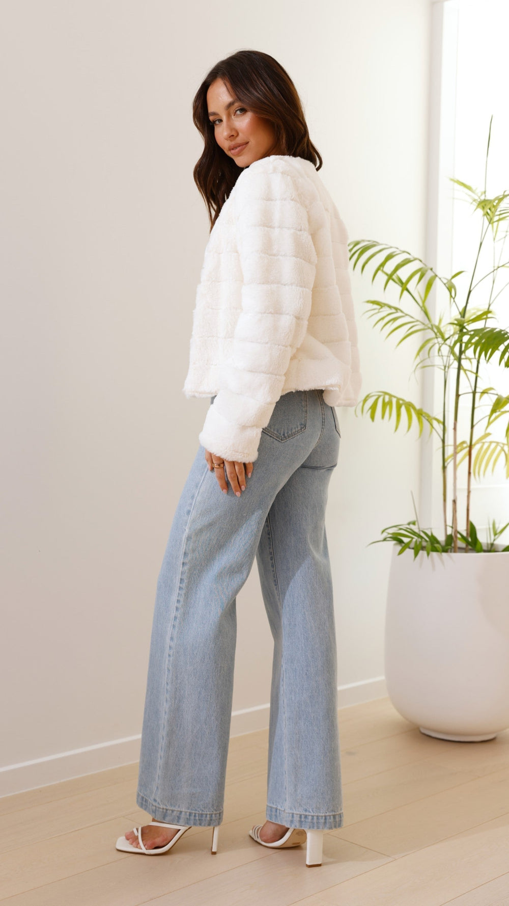 Woman wearing a white puffer jacket and light blue jeans standing indoors with a plant in the background.
