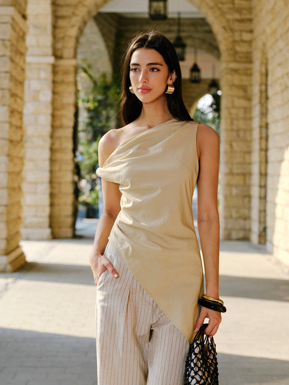 Woman in a beige one-shoulder dress standing in front of stone arches.