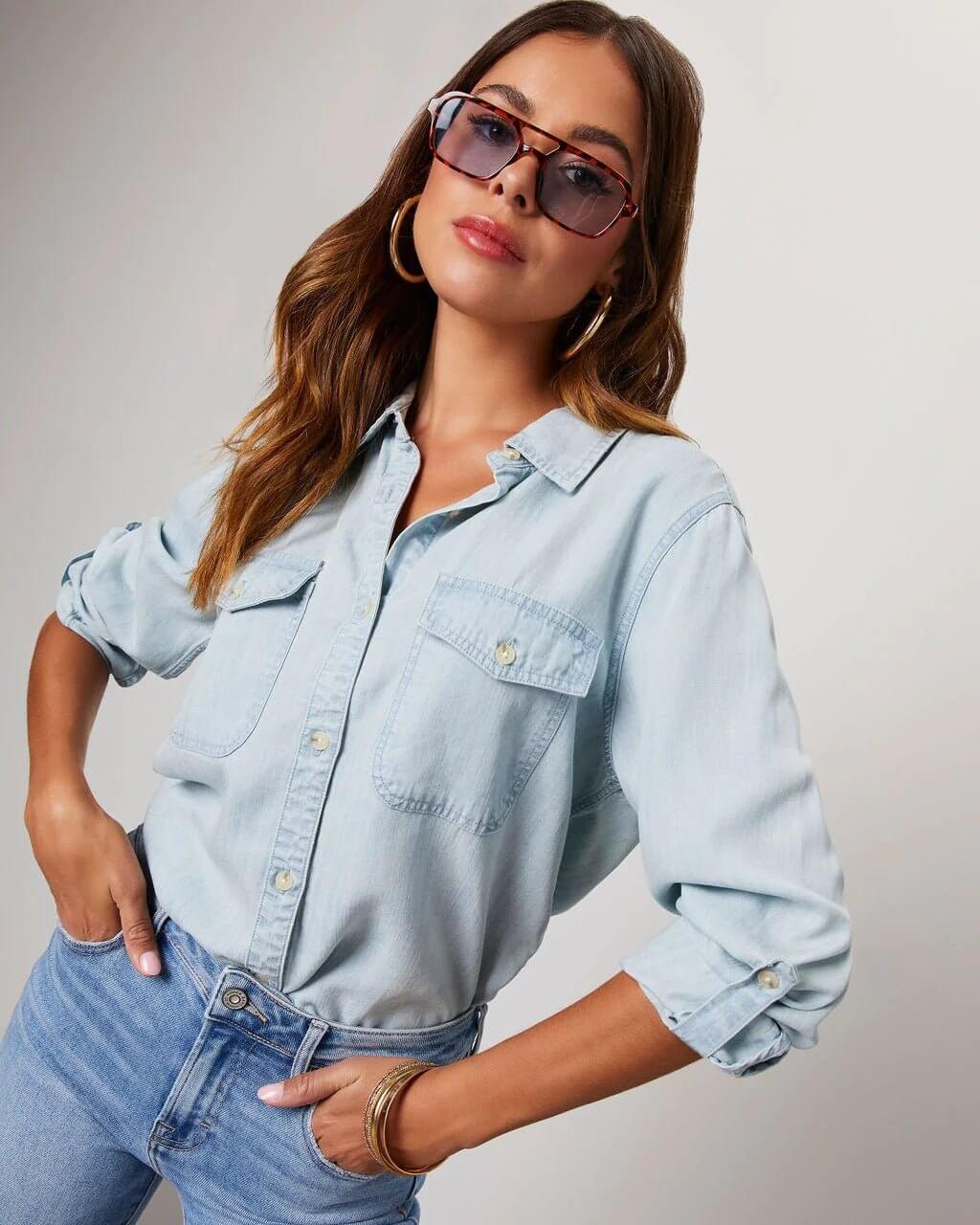 Person wearing a light blue denim shirt and jeans with sunglasses on a neutral background.