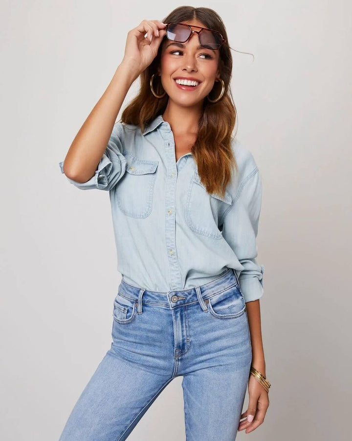 Woman wearing a light blue denim shirt and jeans, smiling and adjusting sunglasses.