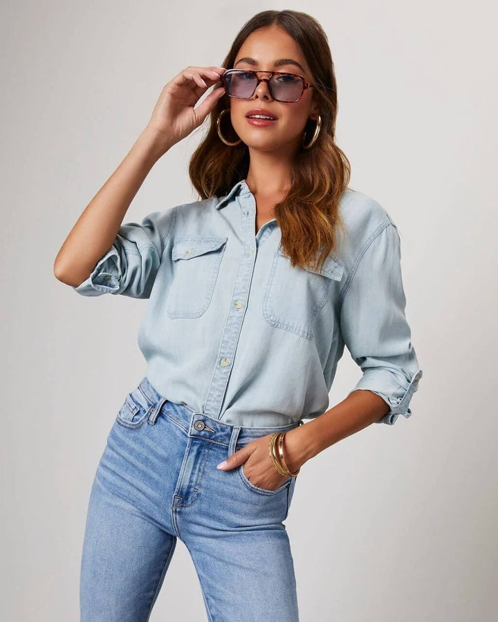 A woman wearing a relaxed fit denim button-up shirt with chest pockets and rolled-up sleeves, paired with jeans and sunglasses.